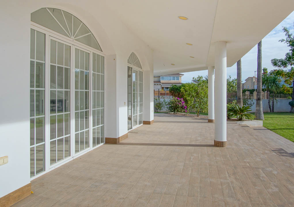 Spacious covered terrace featuring white columns, ceramic flooring, and large arched glass doors overlooking the manicured garden with palm trees.