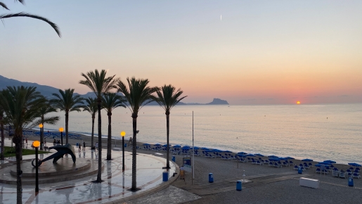 Sunrise view from the nearby promenade featuring palm trees, an anchor sculpture, and a sandy beach with blue umbrellas along the Mediterranean coast.