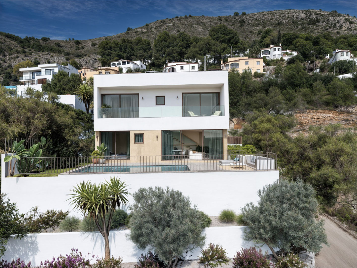 Exterior view of the modern villa featuring a white and beige facade, private pool, large glass sliders, and glass-railed balconies set against a mountain backdrop.