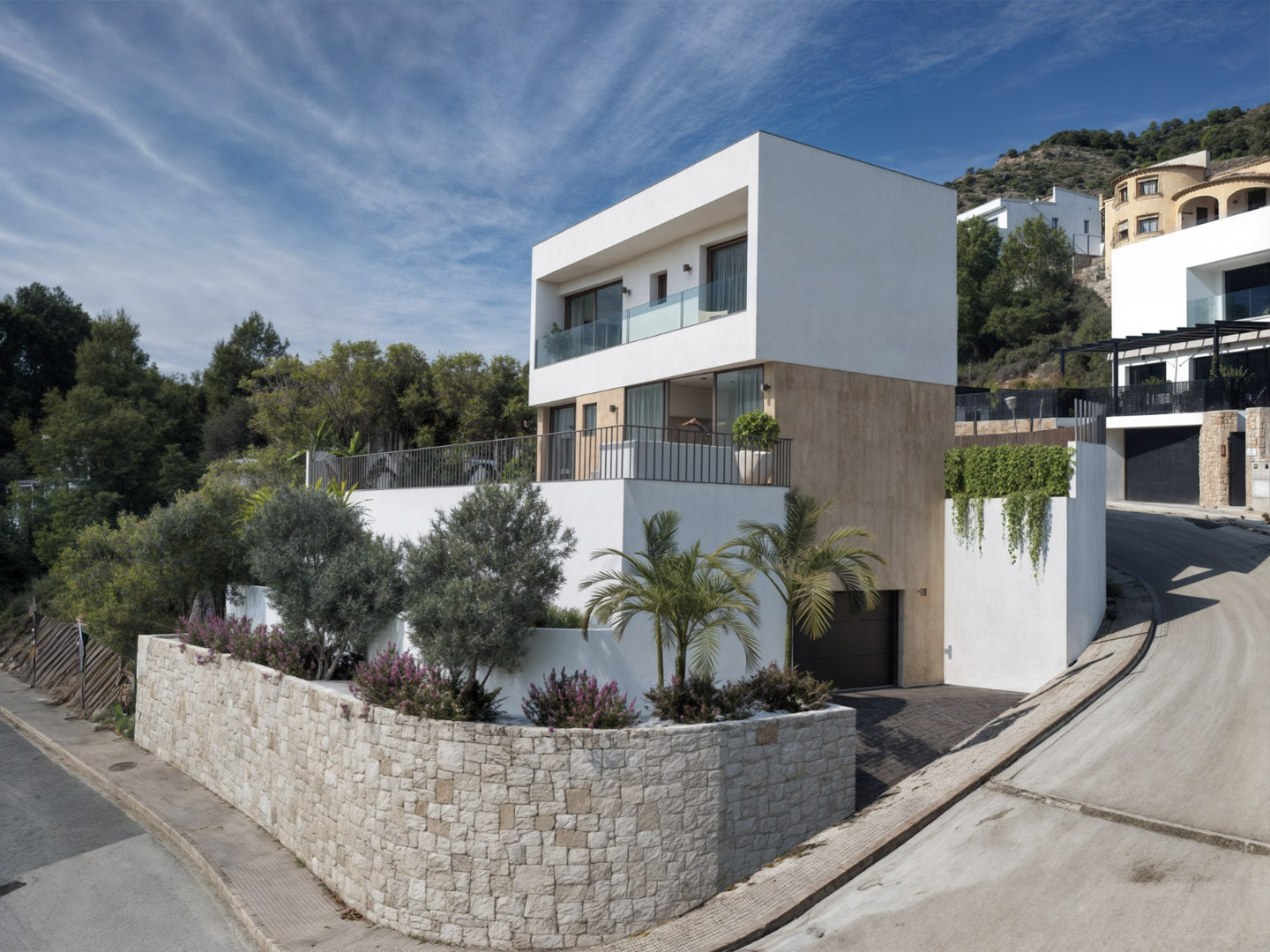 Modern three-story villa featuring a natural stone facade, glass-railed terraces, and a semi-basement garage, set against a Mediterranean hillside landscape.
