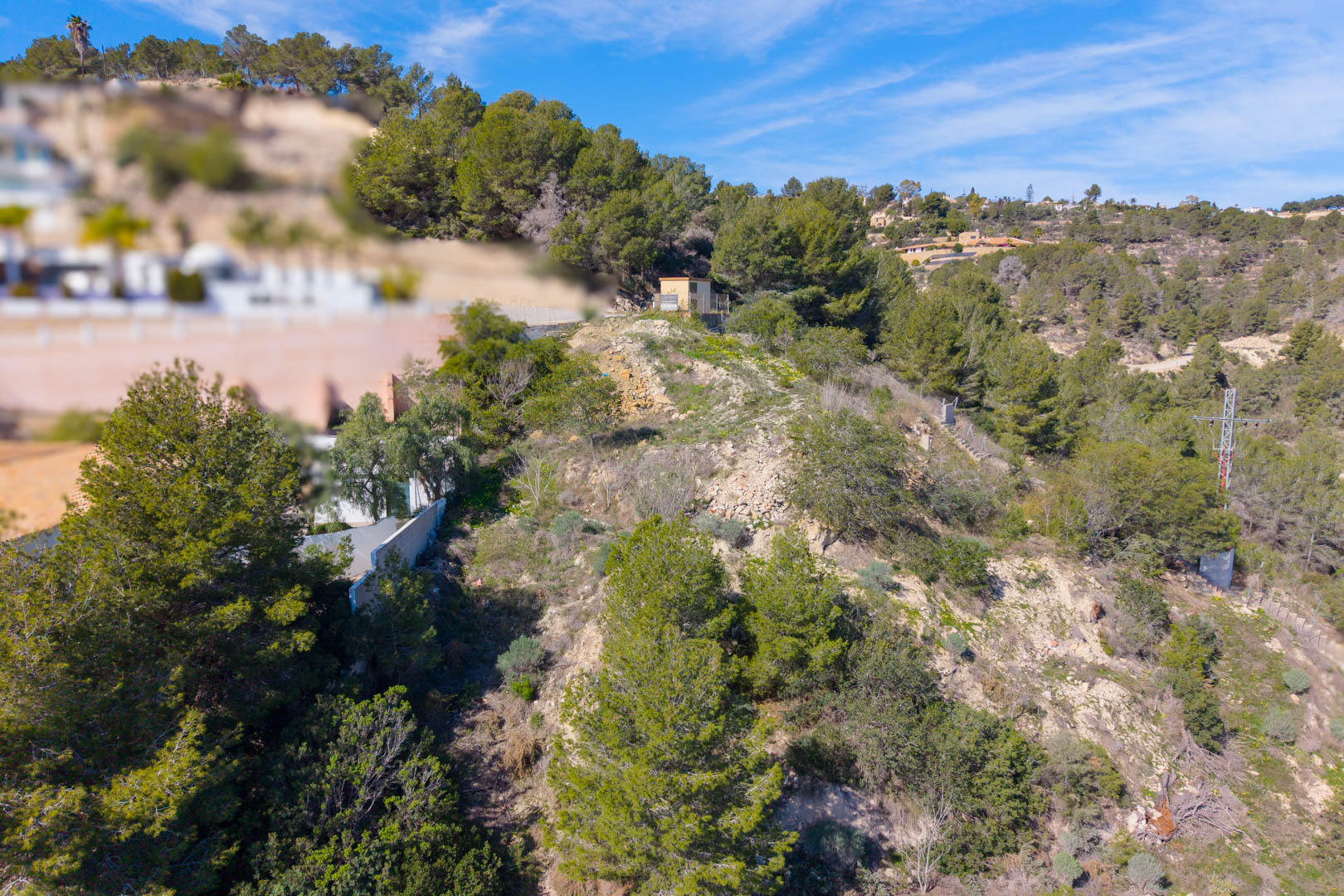 Steep hillside plot in Tossal Los Bancales featuring Mediterranean pines, natural shrubland, and existing utility infrastructure under a clear sky.