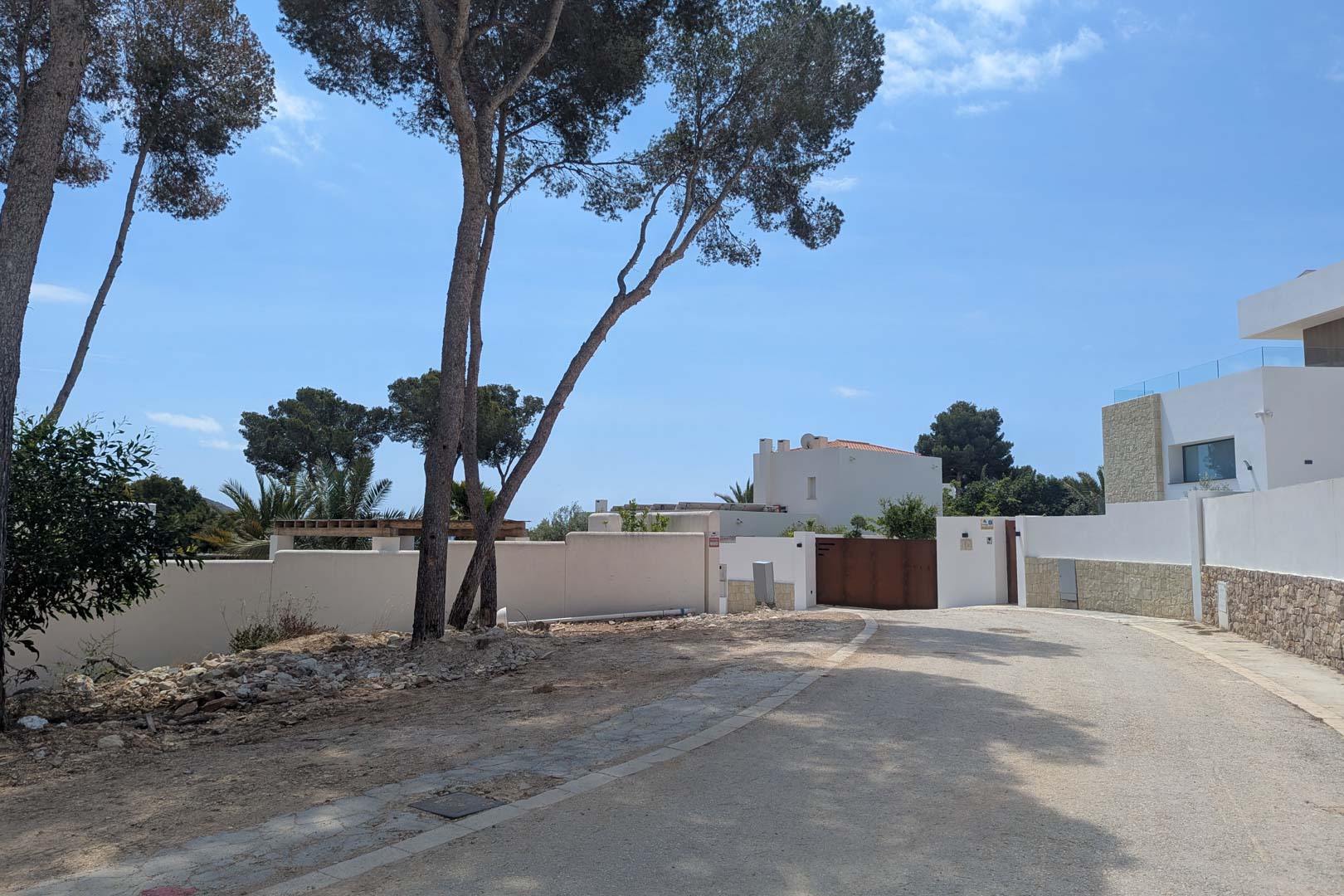 Access to the plot in a Moraira residential area, featuring white perimeter walls, mature pine trees, and a paved road under a clear blue sky.