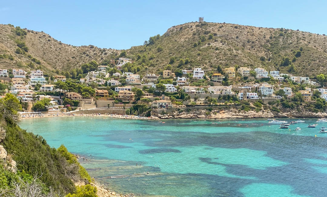Panoramic view of El Portet beach in Moraira, featuring turquoise waters, anchored boats, and white villas nestled along the scenic Mediterranean hillside.