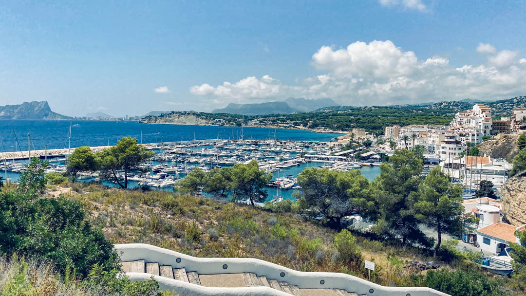 Panoramic view of Moraira marina and the Peñón de Ifach from a Mediterranean-style white staircase surrounded by native coastal vegetation.