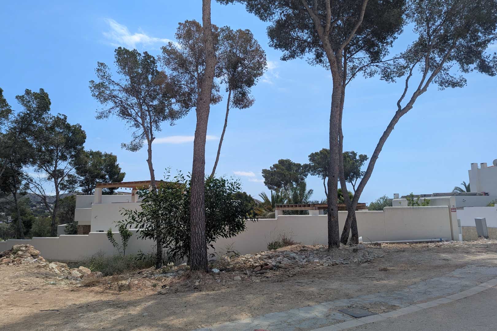 View of the building plot featuring mature pine trees and white perimeter walls, ready for the construction of the Ibizan-style villa in Moraira.