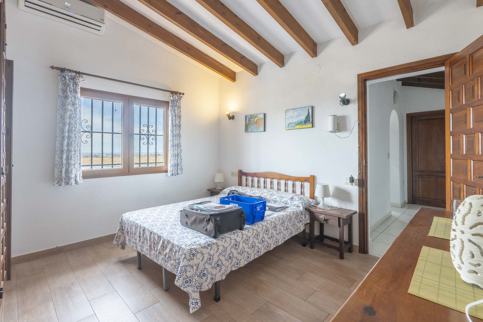Bright bedroom featuring wood beams, a double bed with patterned linens, and an exterior view.