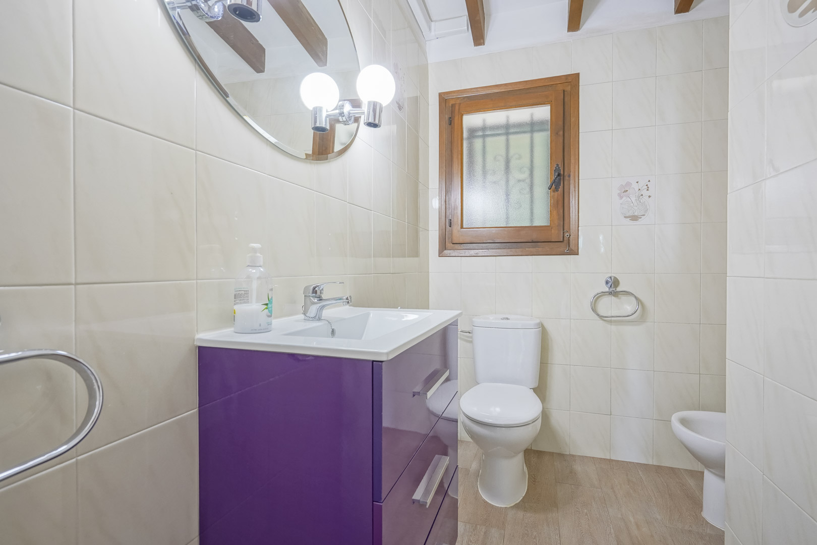 Close-up of a bathroom featuring cream-colored tiles, a purple vanity, and a wood-framed window.
