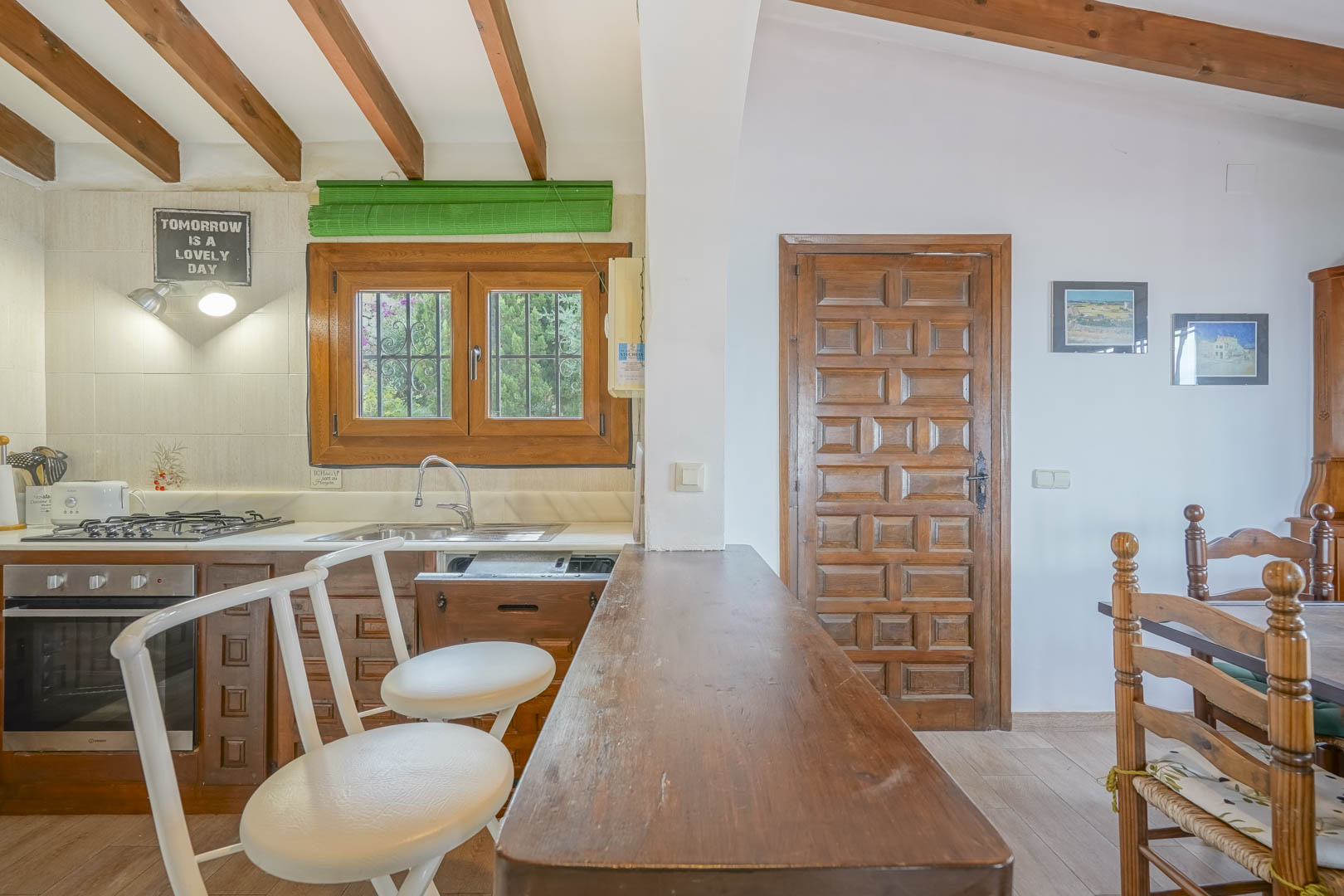 Close-up of the open kitchen featuring a wooden bar, white stools, and exposed ceiling beams.