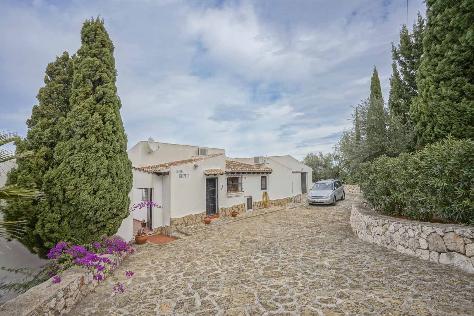Front view of the villa with stone driveway, gardens, and white facade with rustic details.