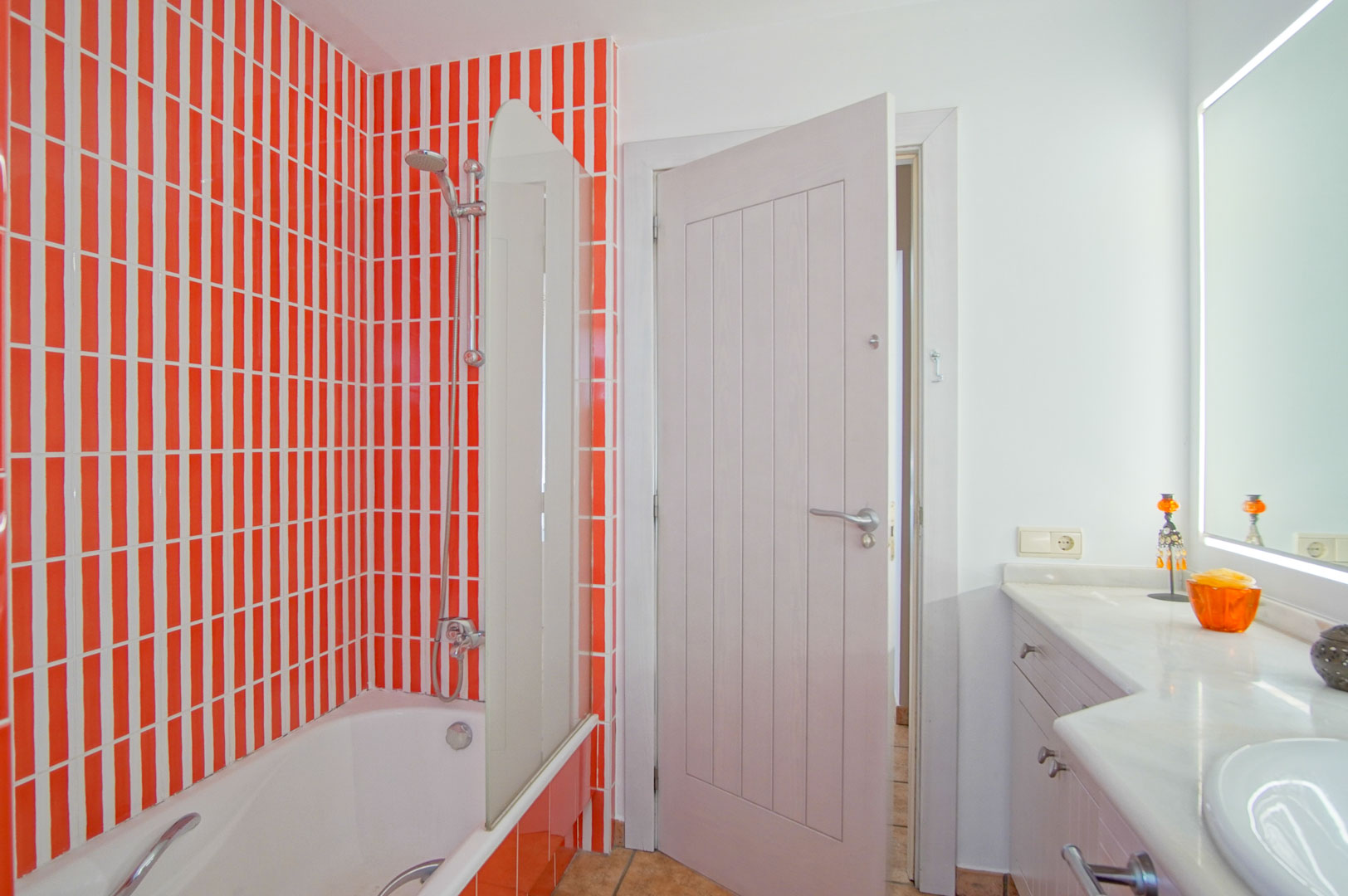 Bathroom featuring a bathtub with a glass screen, distinctive orange and white vertical tiling, and a white vanity with a marble countertop and backlit mirror.