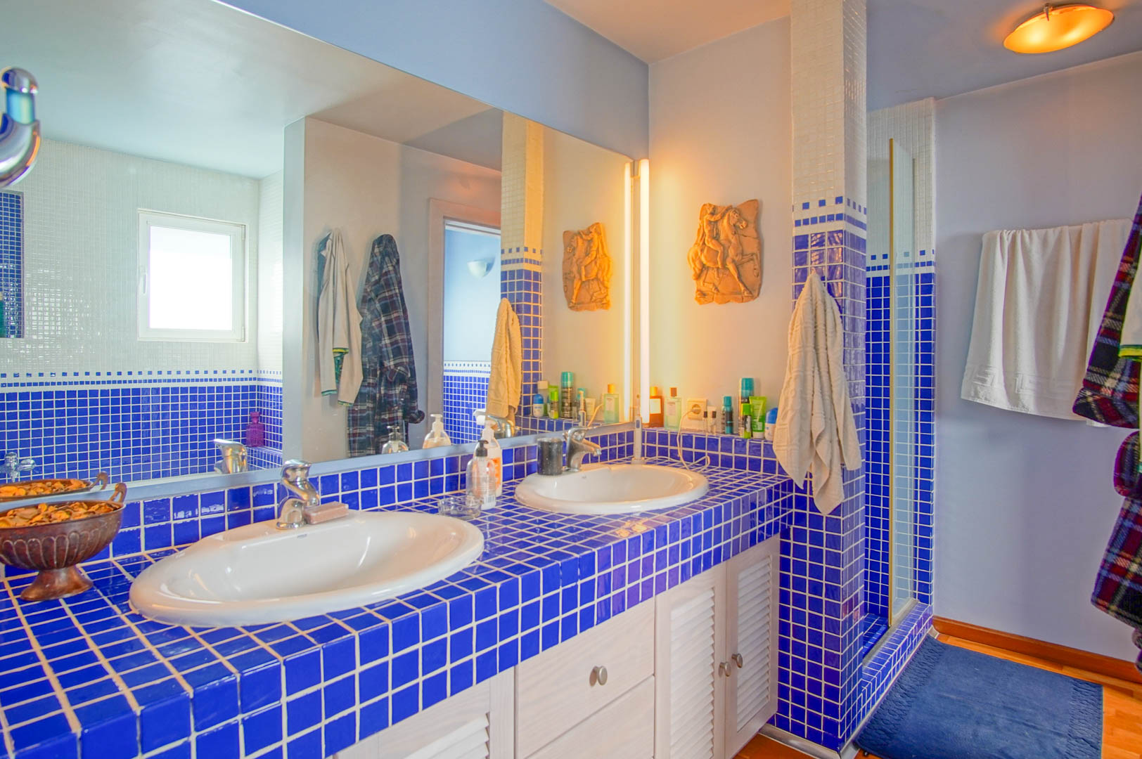 Bathroom featuring a blue-tiled double vanity with white louvered cabinets. A large wall mirror reflects the integrated shower area and warm ambient lighting.