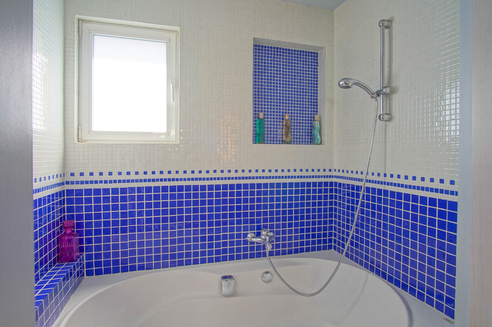Bathroom featuring an oval bathtub and cobalt blue and white mosaic tiling. It includes a built-in storage niche and a frosted window for natural ventilation and light.