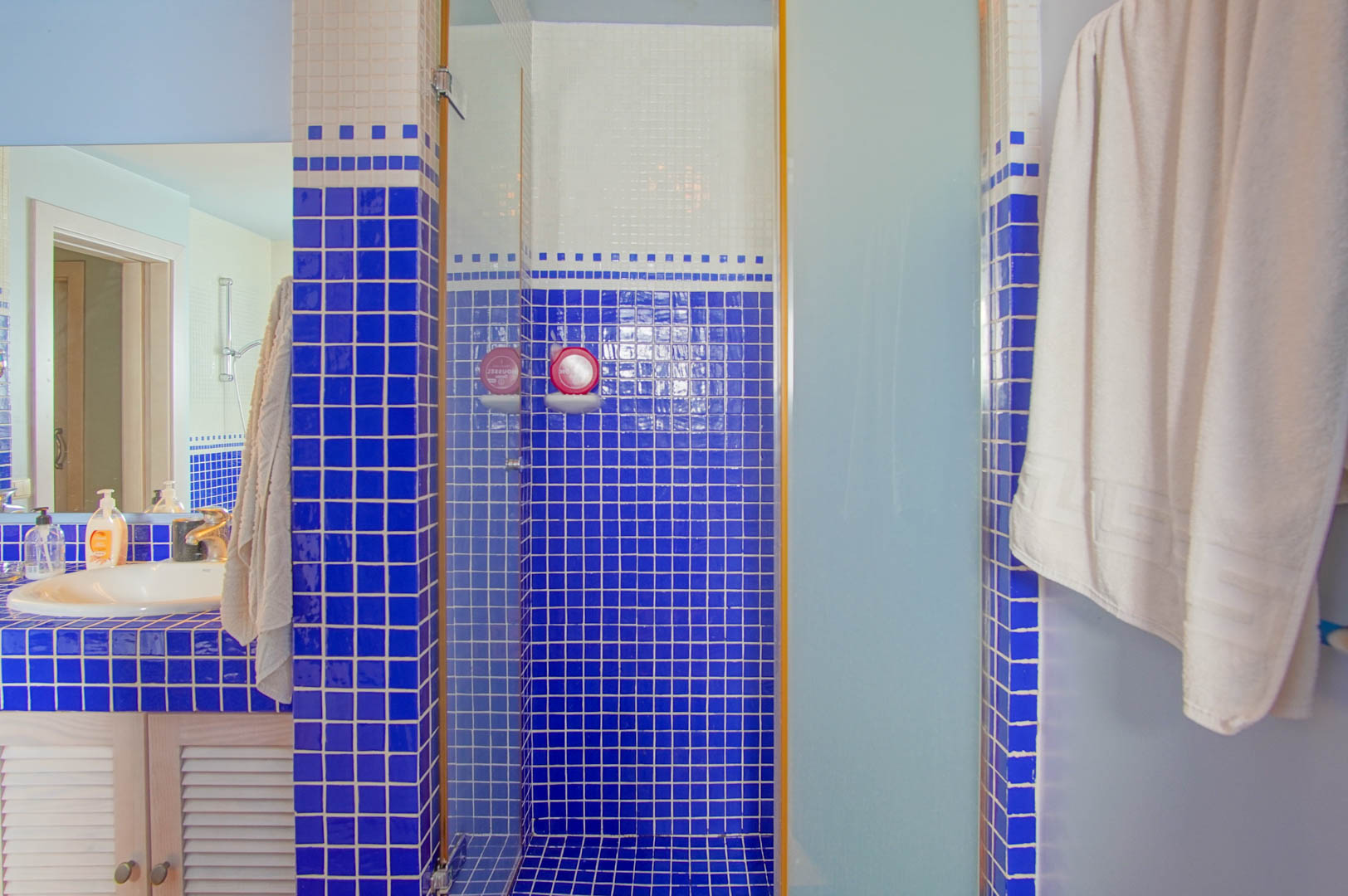 Bathroom featuring blue and white mosaic tiling, a walk-in shower with glass enclosure, and a vanity with a white basin and louvered wooden doors.