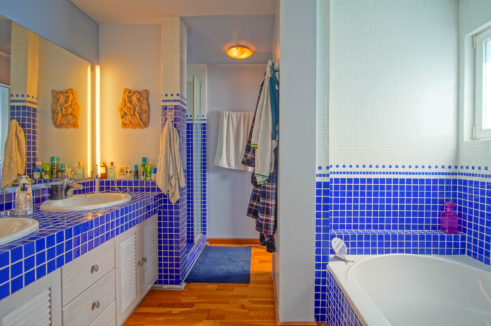 Bathroom featuring blue and white mosaic tiling, double vanity with louvered cabinets, wooden flooring, and a built-in bathtub illuminated by natural and recessed light.