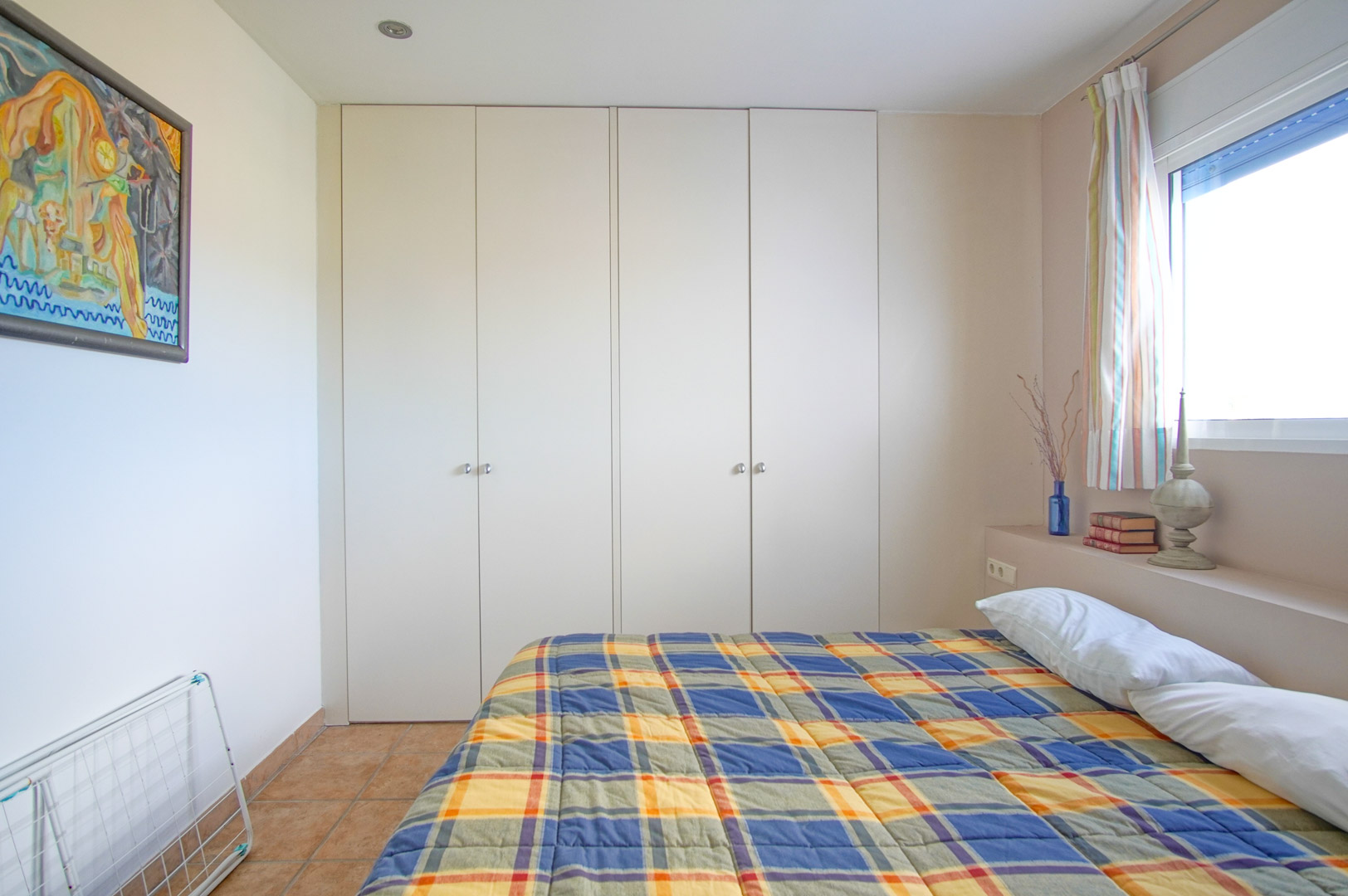 Bright bedroom featuring large white built-in wardrobes, rustic tiled flooring, and a side window providing ample natural light to the space.