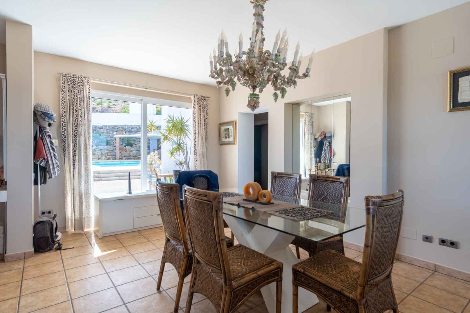 Bright dining area featuring a glass-top table, wicker chairs, and an ornate chandelier, offering direct views and access to the outdoor pool area through large windows.