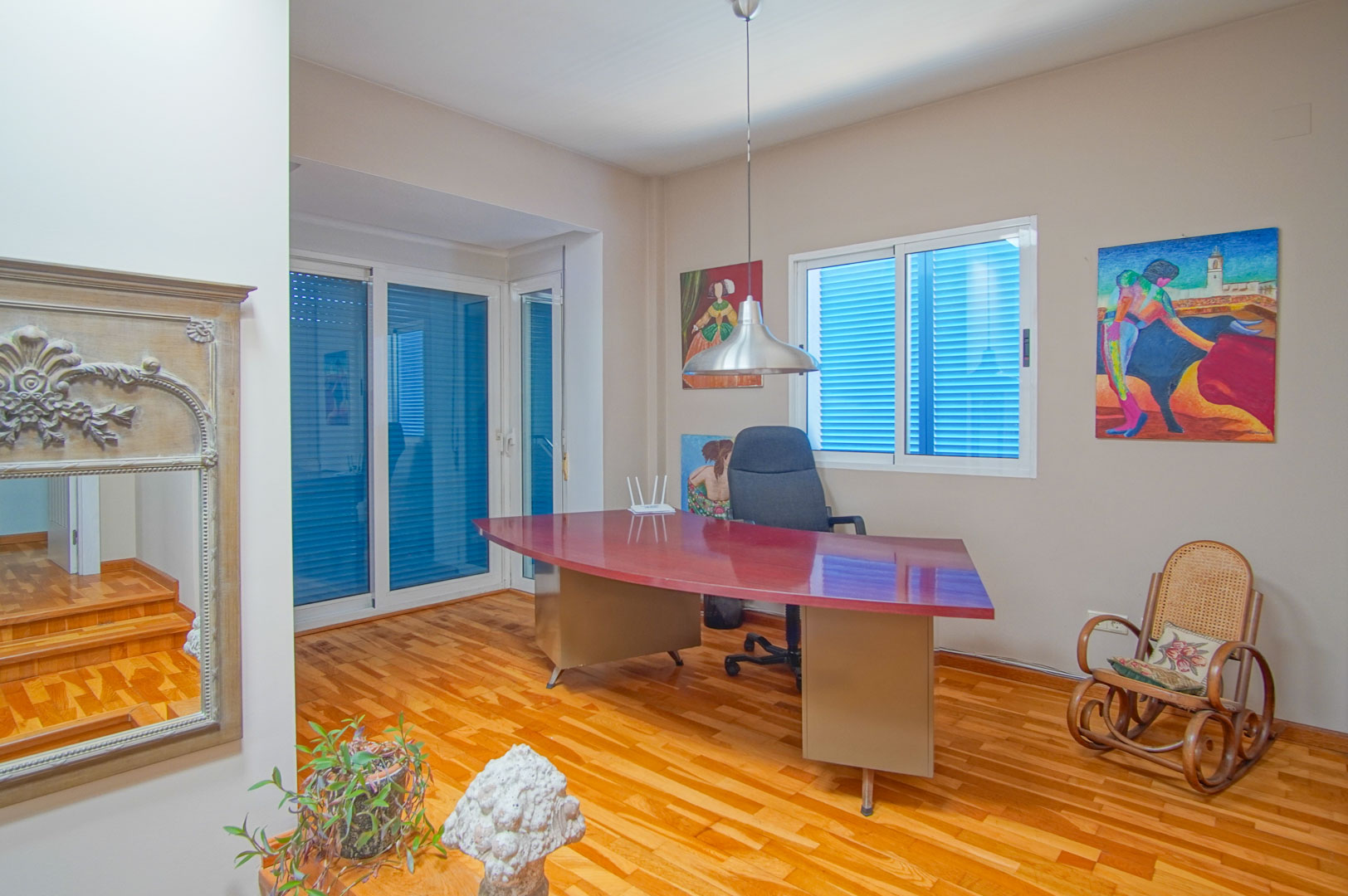 Bright home office featuring parquet flooring, a large curved burgundy desk, and windows with traditional blue shutters providing natural light.