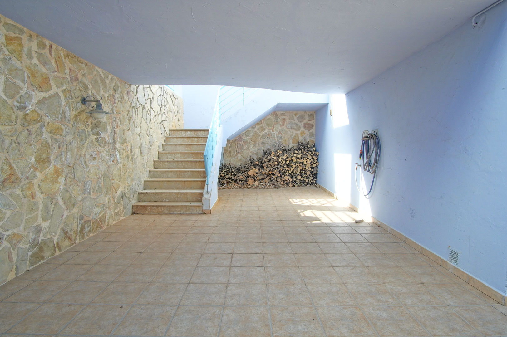 Covered patio featuring a natural stone wall, ceramic tiling, and a dedicated firewood storage space beneath the exterior staircase.