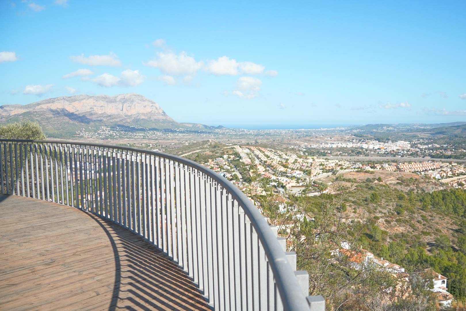 Curved wooden deck terrace with a metal railing featuring panoramic views of the valley, surrounding mountains, and the Mediterranean coastline.