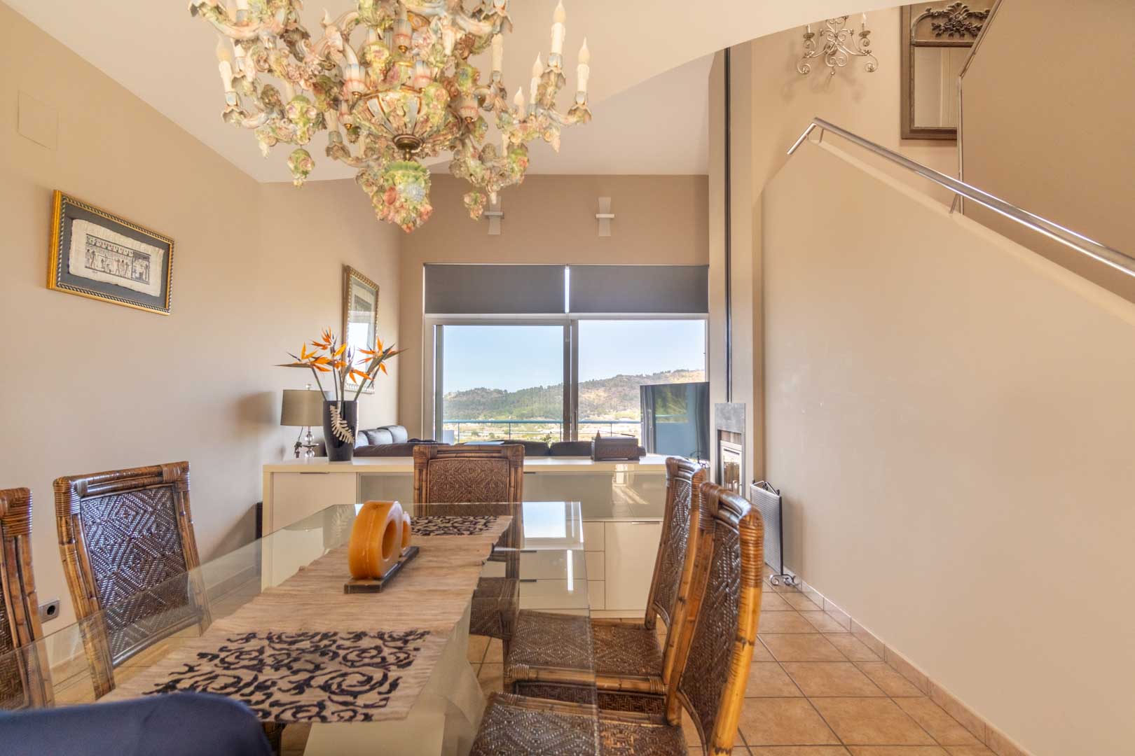 Dining area featuring a glass table and woven chairs, highlighted by an ornate chandelier and panoramic mountain views through large windows.