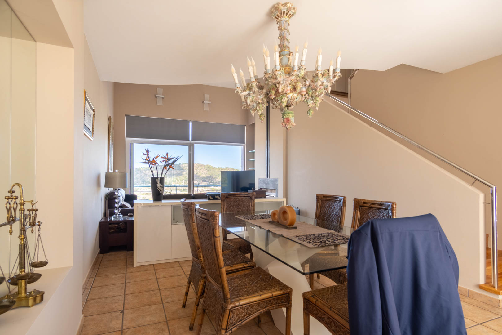 Dining area featuring a glass-top table, wicker chairs, and an ornate porcelain chandelier. A large window offers mountain views next to the staircase with metal railings.
