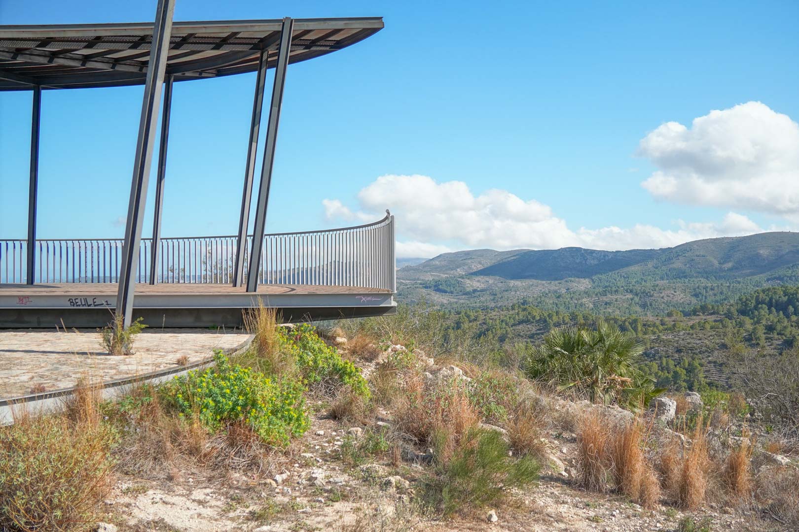 Elevated observation deck featuring a steel frame and metal railing, overlooking the expansive mountain range and lush Mediterranean valley scenery.