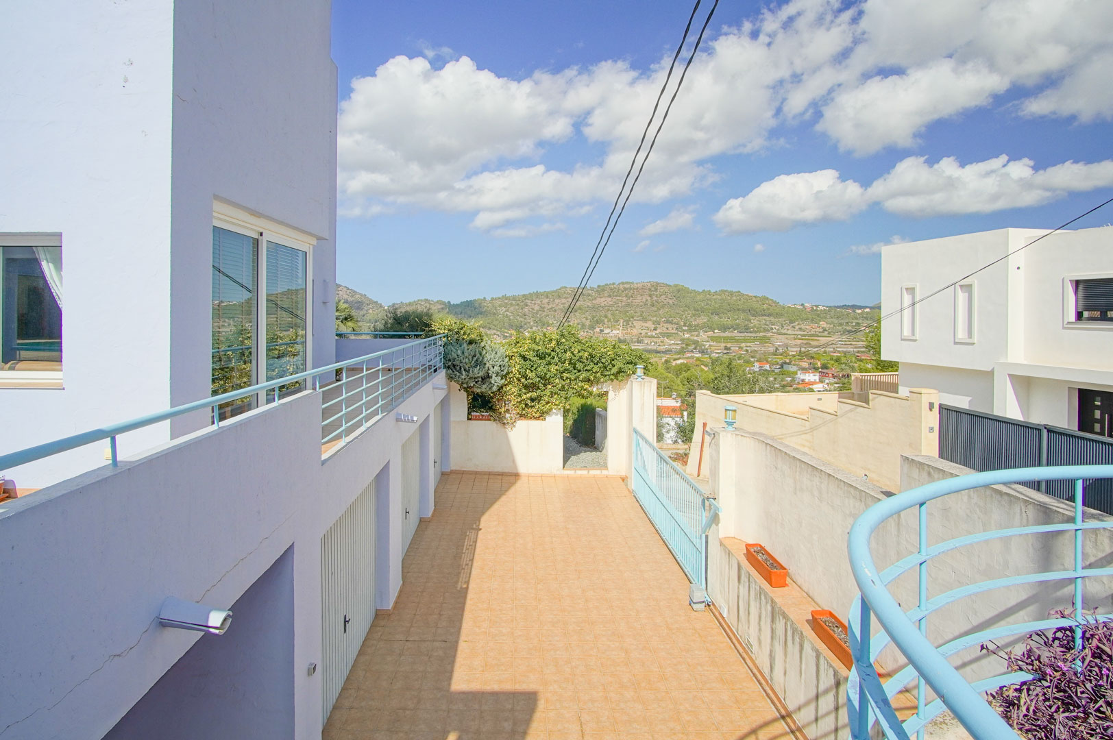 Exterior view of the driveway featuring three private garages, terracotta tiling, and blue railings, overlooking the scenic mountain landscape.