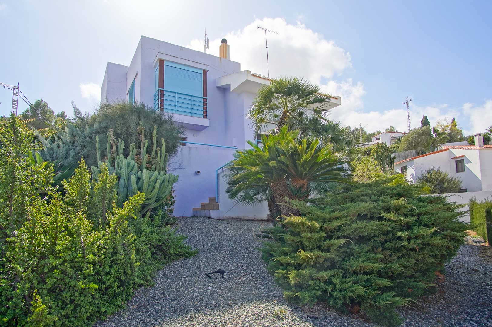 Exterior view of the lavender-toned facade, integrated into a Mediterranean garden with cacti, palm trees, and shrubs on gravel terrain.