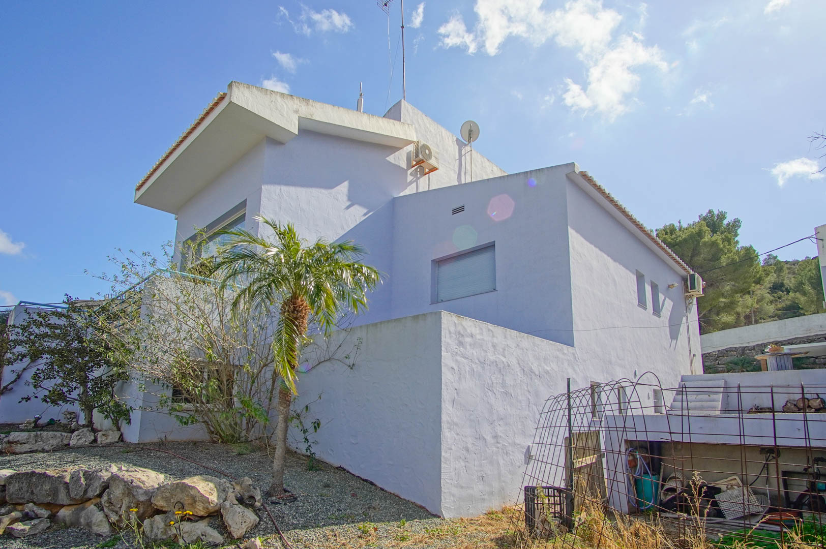 Exterior view of the white villa's facade on a sloping plot with gravel landscaping, rocks, and a palm tree, showcasing its multi-level architectural design.