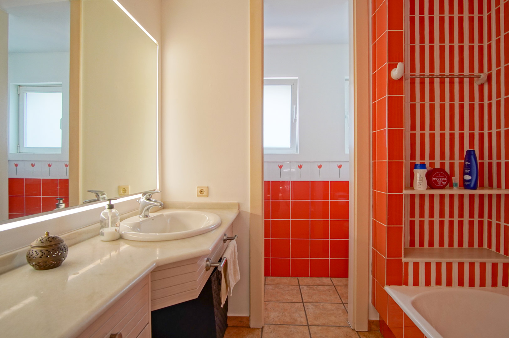 Functional bathroom featuring a marble countertop, backlit mirror, and vibrant orange tiling. Includes a bathtub area with integrated shelving and natural light.