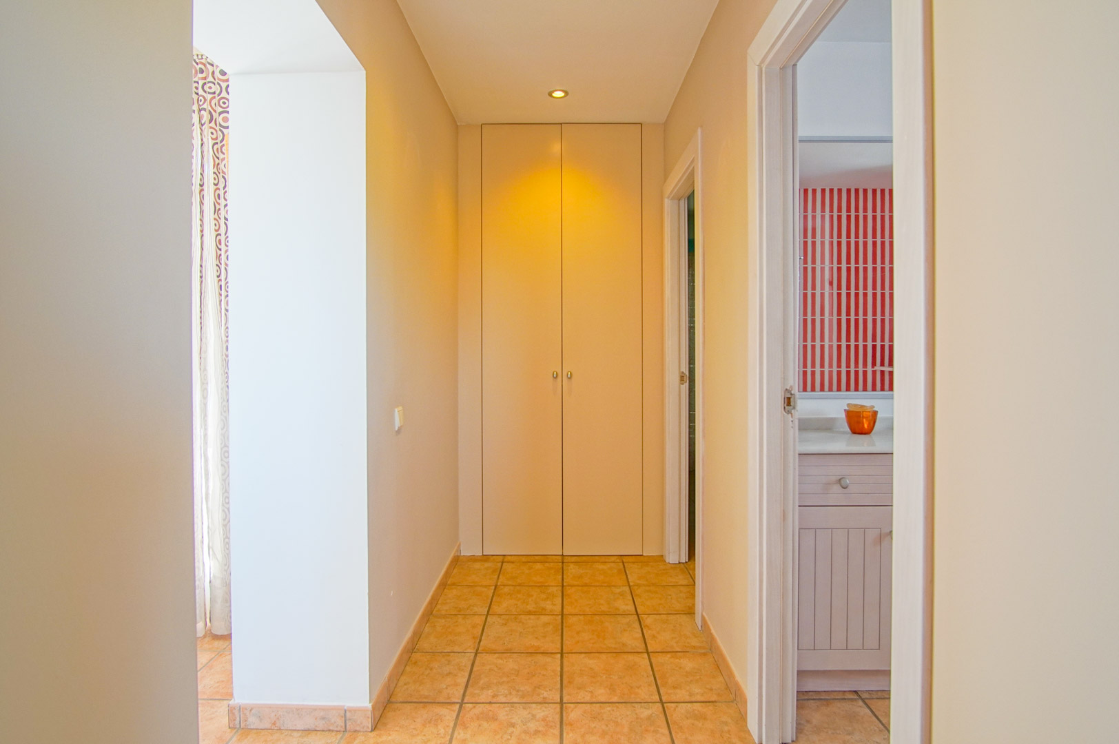 Hallway featuring terracotta-style floor tiles, a built-in floor-to-ceiling wardrobe, and a view into a bathroom with distinctive red and white striped tiling.