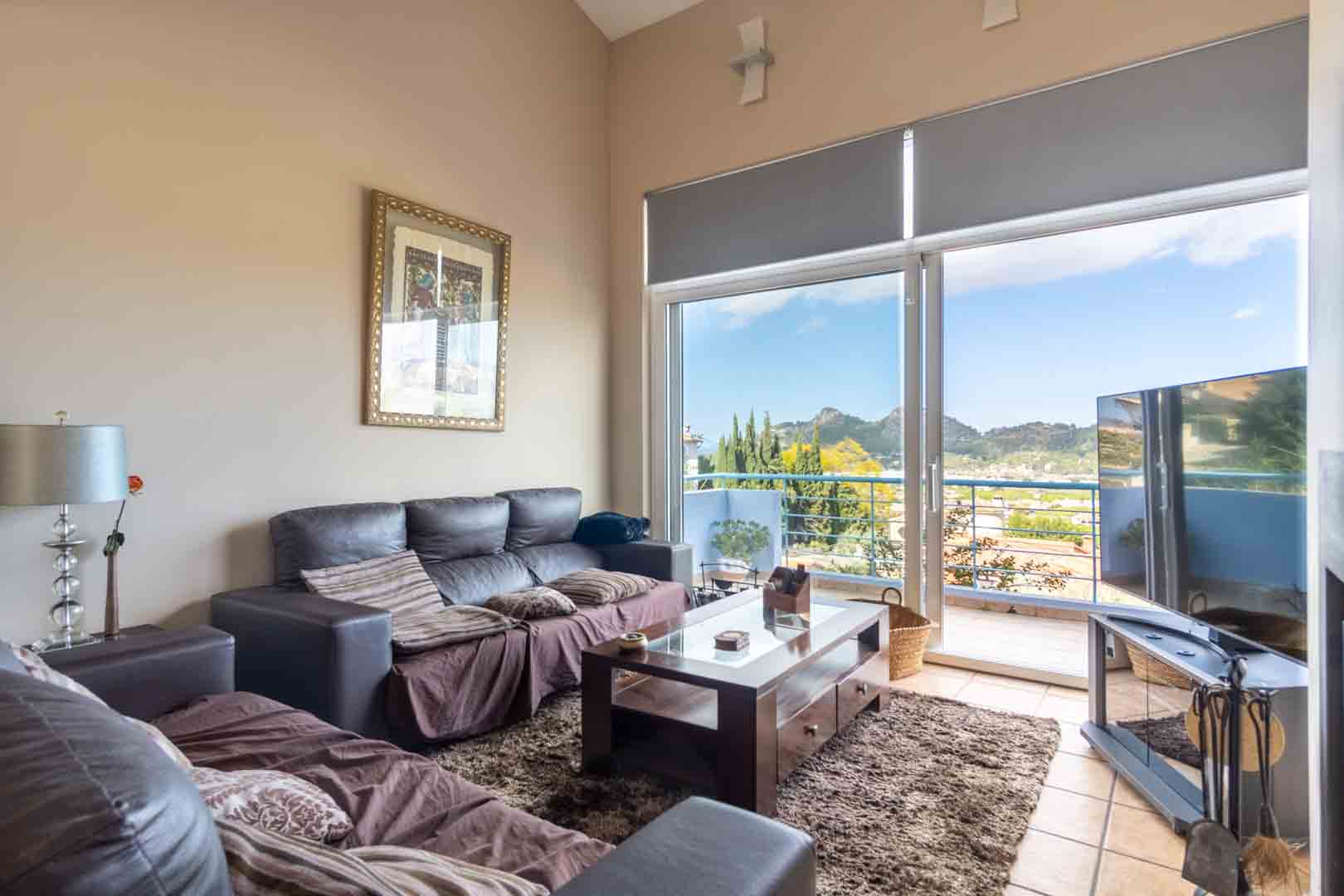 Living room featuring high ceilings, dark sofas, and a wooden coffee table. Large sliding glass doors open to a balcony with expansive mountain and valley views.