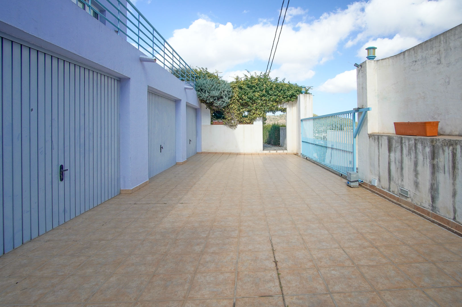 Lower entrance featuring three private garages with light blue doors, a spacious tiled driveway, and a secure sliding metal gate under the upper terrace.
