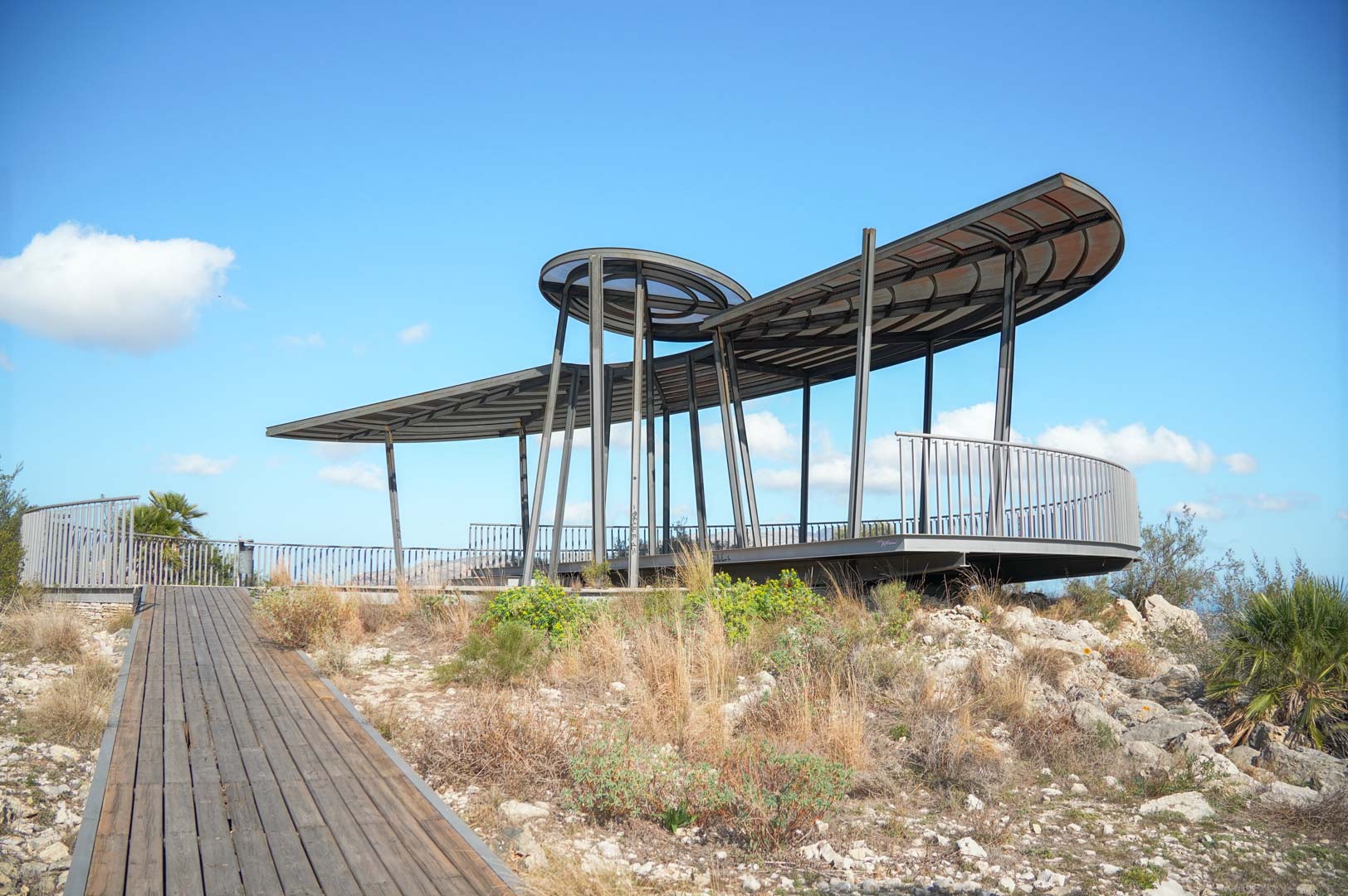 Modern architectural viewpoint featuring a curved metal roof and steel structure, accessed by a wooden walkway amidst natural Mediterranean vegetation.