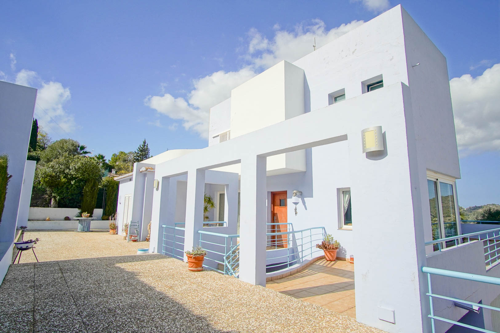 Modern white facade with light blue accents, featuring a spacious aggregate terrace and a wooden entrance door framed by minimalist metal railings.