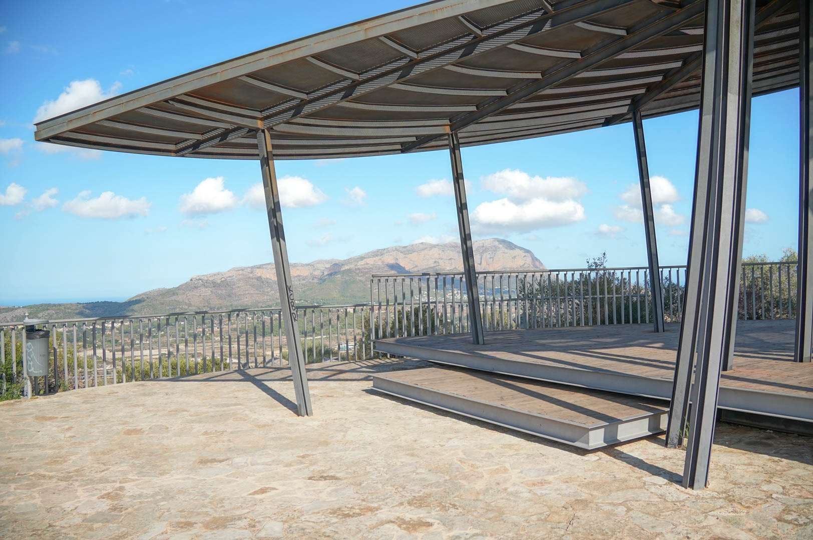 Outdoor viewpoint featuring a modern steel structure and wooden decking, providing panoramic mountain and valley views under a clear sky.