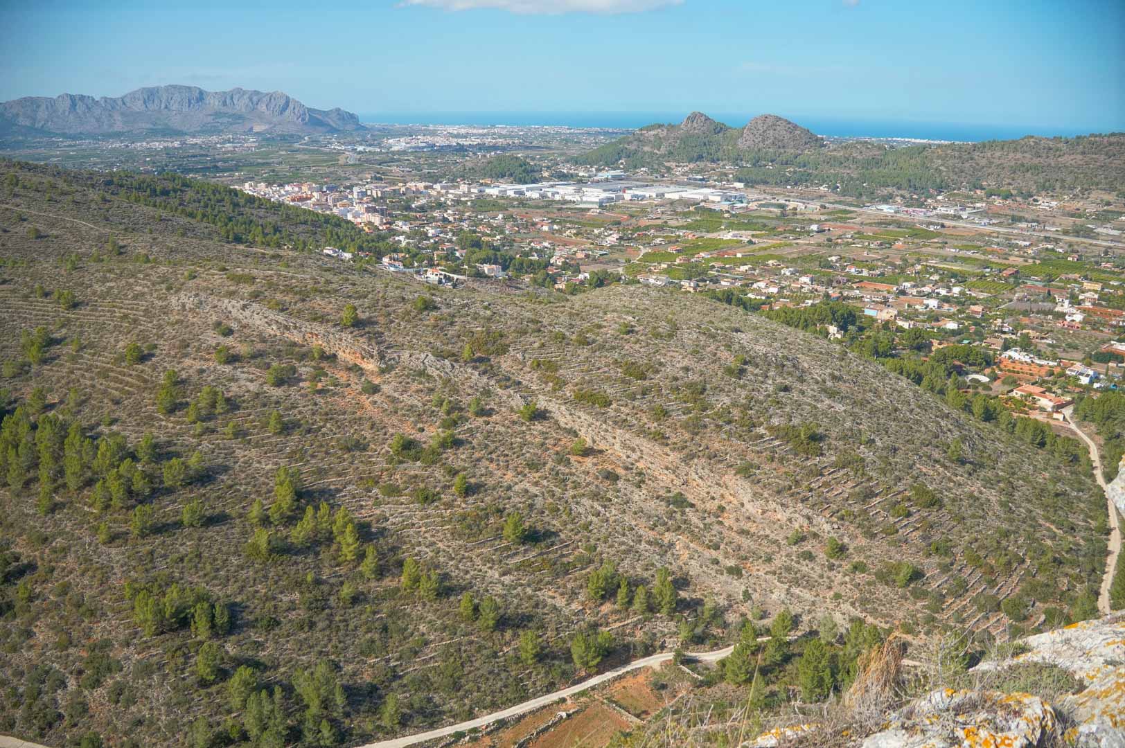 Panoramic elevated view overlooking the Pedreguer valley, featuring rugged mountain ridges and the Mediterranean Sea visible on the distant horizon.