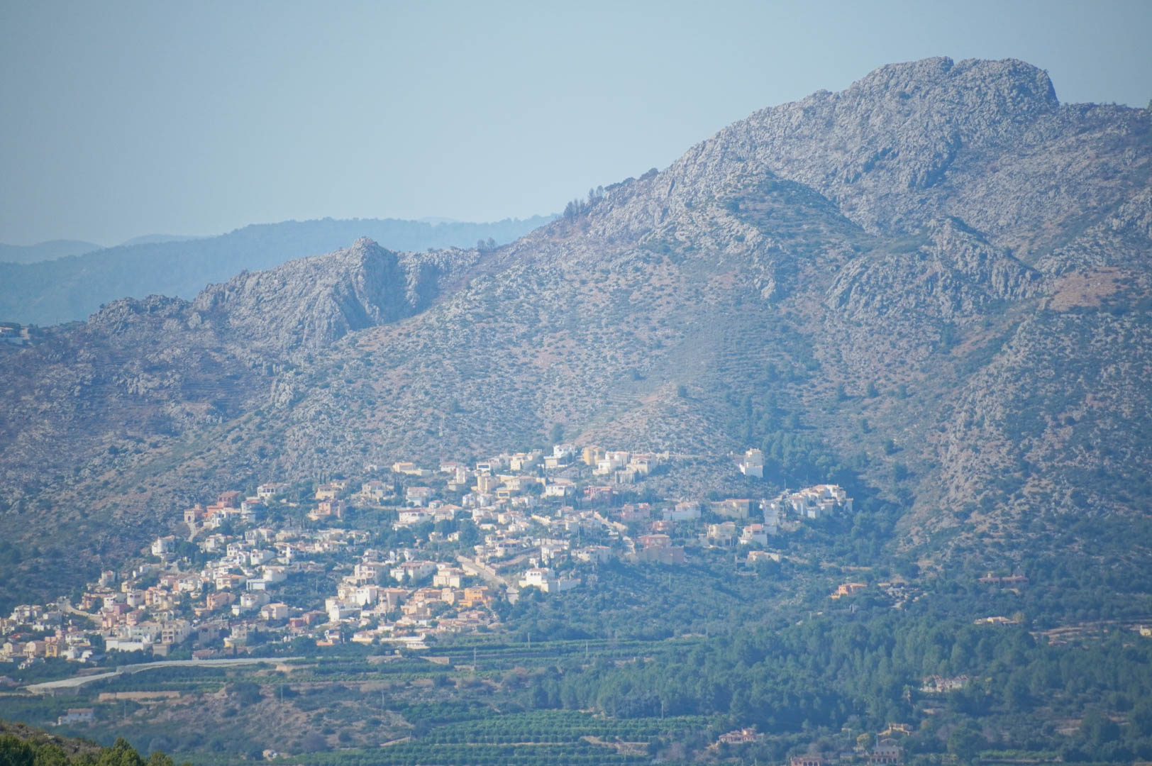 Panoramic view of the rugged mountain range and valley, showcasing a residential cluster on the hillside above lush green agricultural fields.