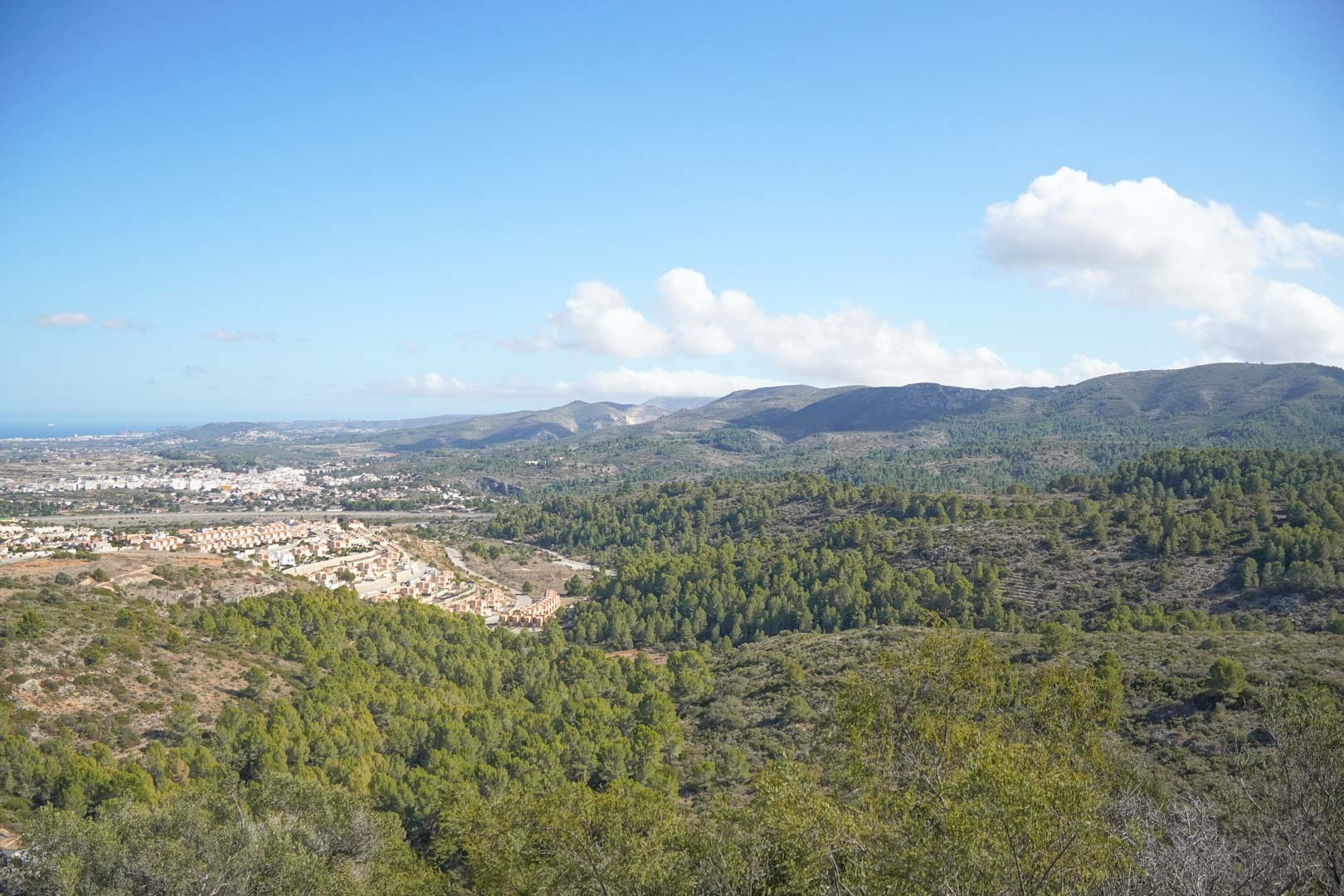 Panoramic views overlooking the Pedreguer valley, featuring pine-covered mountains, residential areas, and the Mediterranean Sea on the horizon under a clear sky.