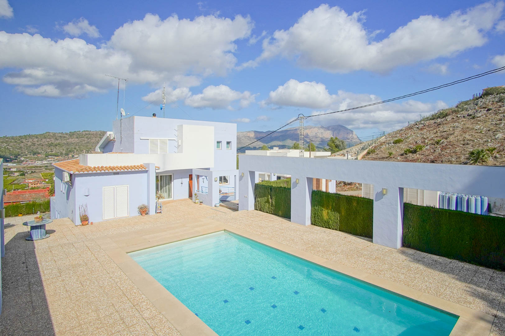 Rectangular swimming pool and stone-paved terrace set against the villa's lavender facade, offering clear views of the surrounding mountains.