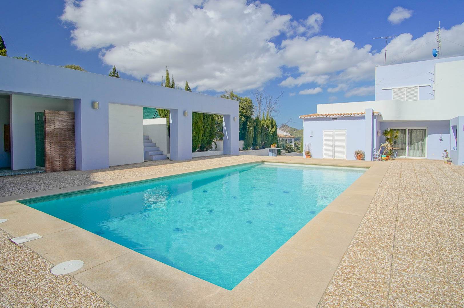 Rectangular swimming pool featuring a stone and gravel terrace, framed by lavender-toned architectural walls and Mediterranean vegetation.