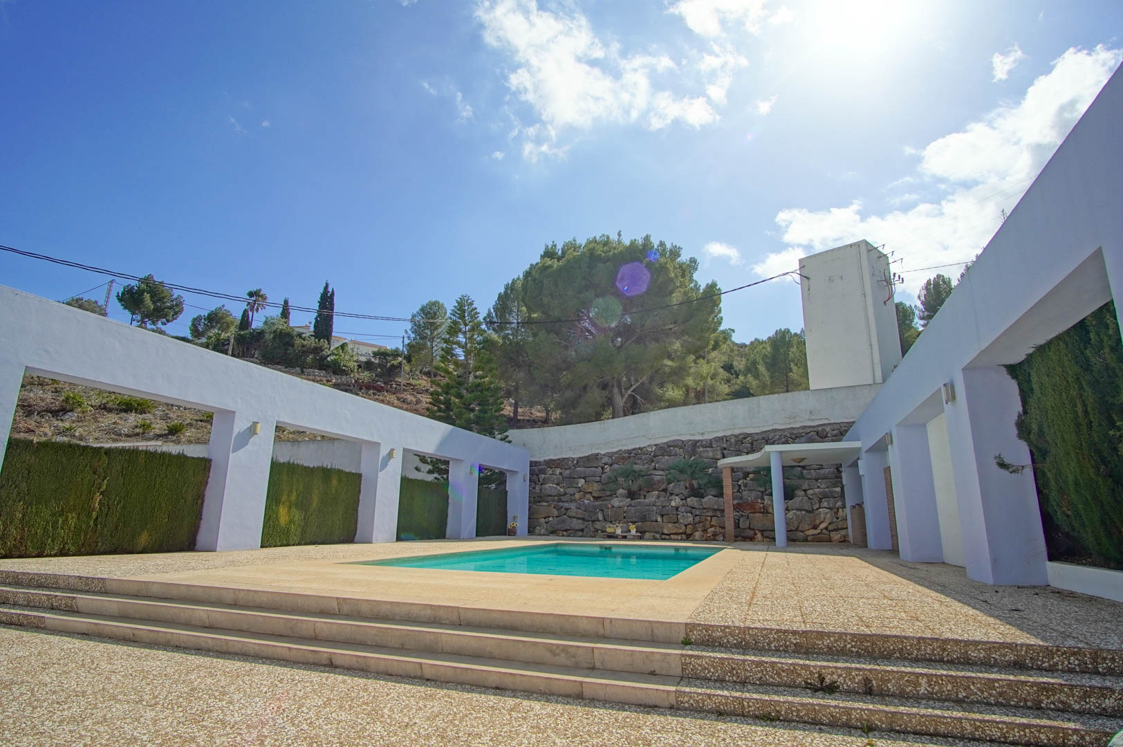 Rectangular swimming pool set within a spacious aggregate terrace, featuring white architectural arches and a backdrop of natural stone walls and pine trees.
