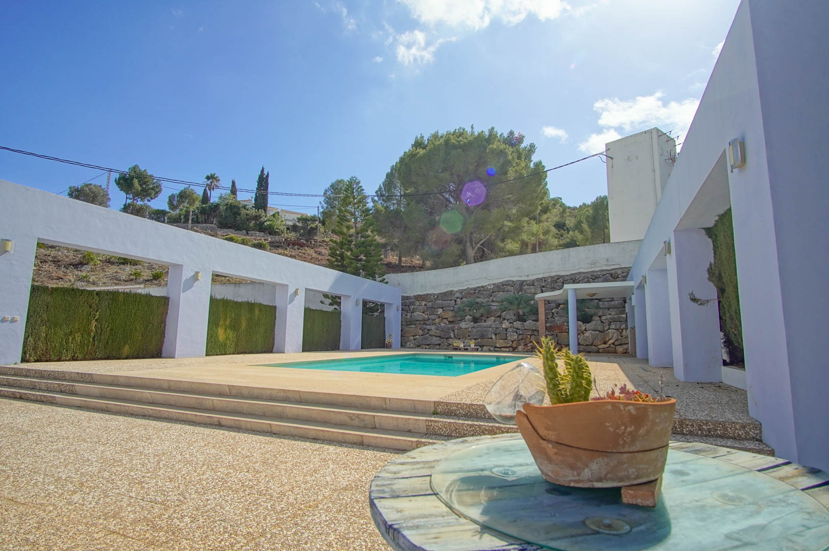 Rectangular swimming pool set within a spacious aggregate terrace, framed by white architectural pillars and a natural stone retaining wall.