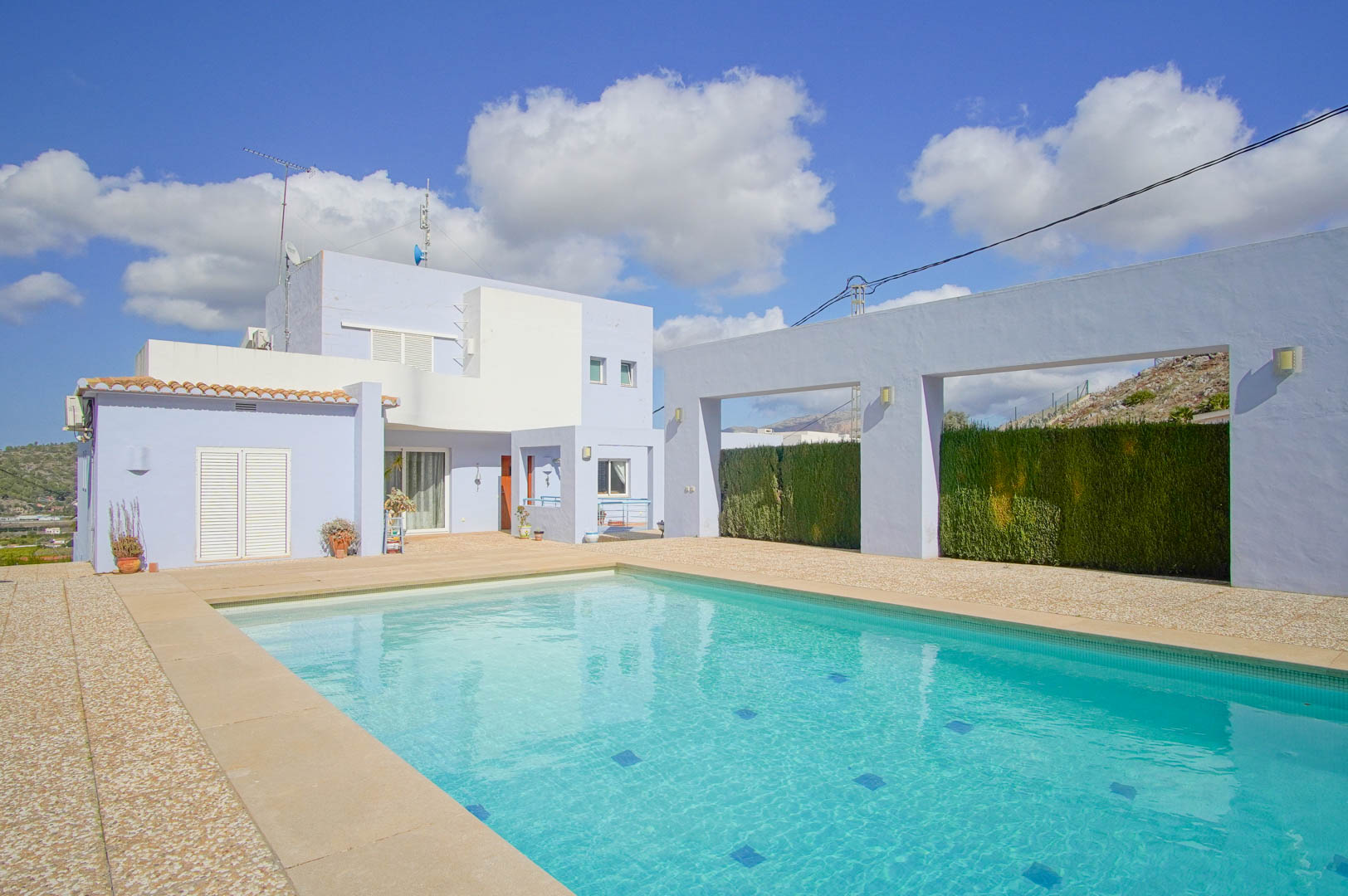 Rectangular swimming pool set within a spacious stone terrace, featuring the villa's modern blue and white facade and architectural privacy walls.