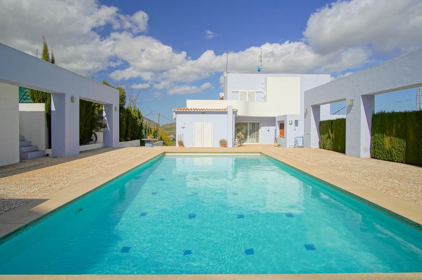 Rectangular swimming pool with turquoise mosaic tiles, framed by modern concrete structures and a spacious gravel terrace leading to the main villa.
