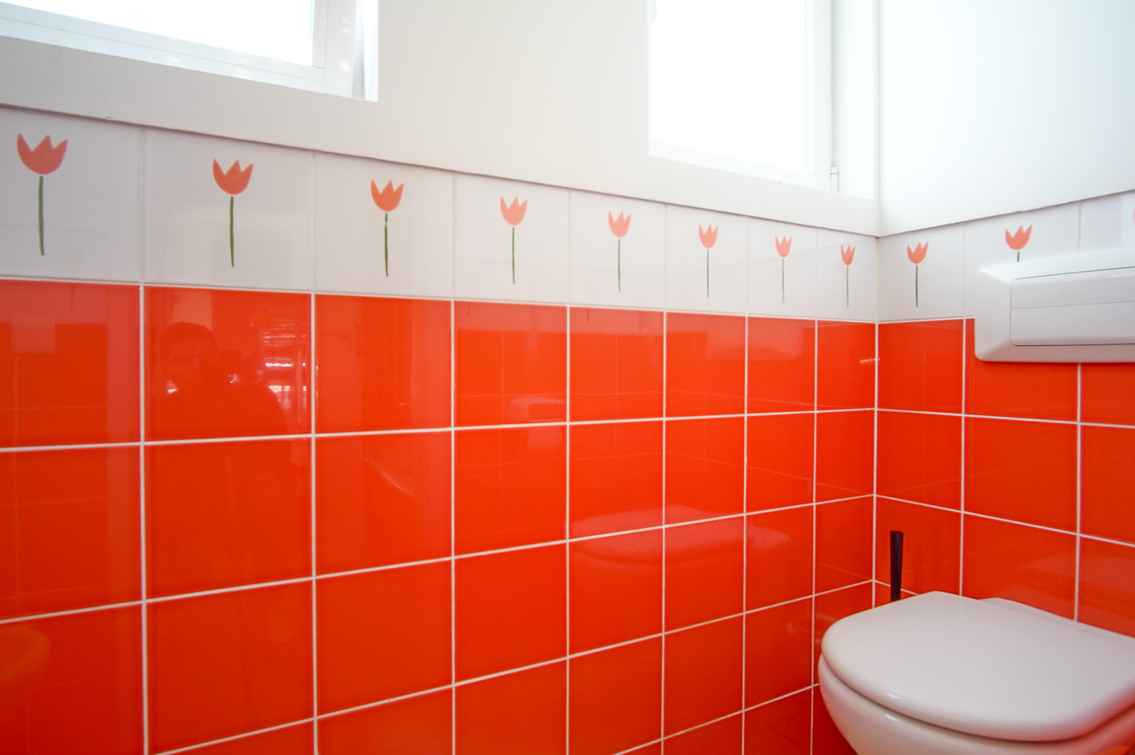 This bathroom features vibrant orange wall tiles and a decorative border with tulip motifs. It includes a wall-hung toilet and benefits from ample natural light.