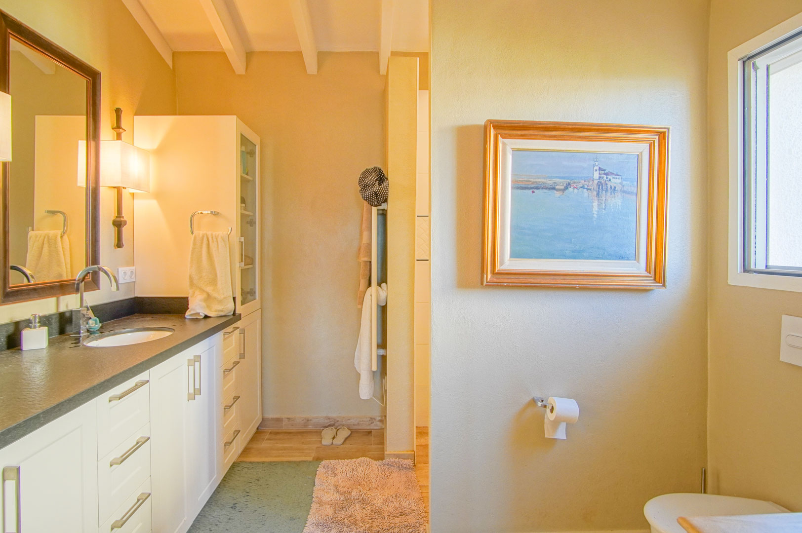 Bathroom featuring a dark countertop, integrated sink, and exposed wooden ceiling beams. It includes white cabinetry, warm wall lighting, and natural light from a side window.