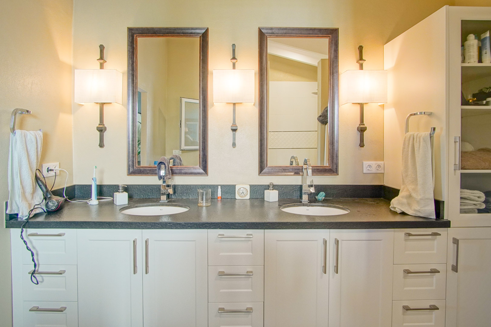 Bathroom featuring a double vanity with dark stone countertops and white cabinetry. Two framed mirrors are flanked by three elegant wall sconces for balanced lighting.