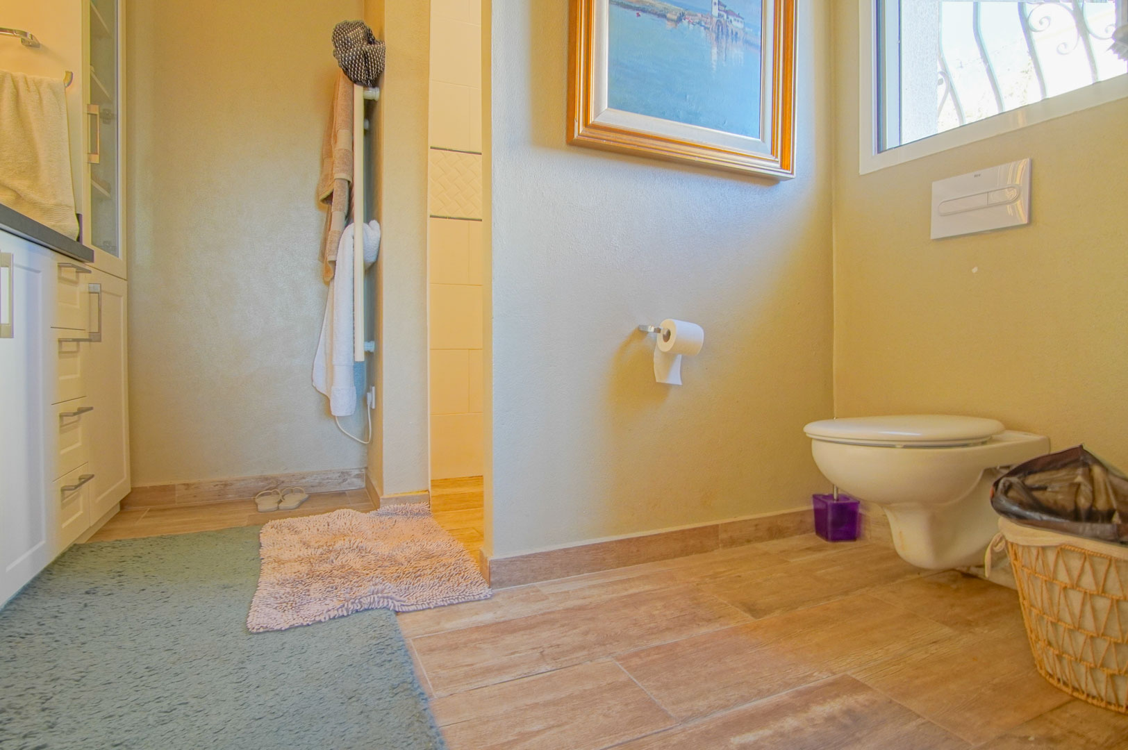 Bathroom featuring a wall-hung toilet, wood-effect ceramic flooring, and white cabinetry. Includes a towel radiator and a glimpse of the tiled walk-in shower.