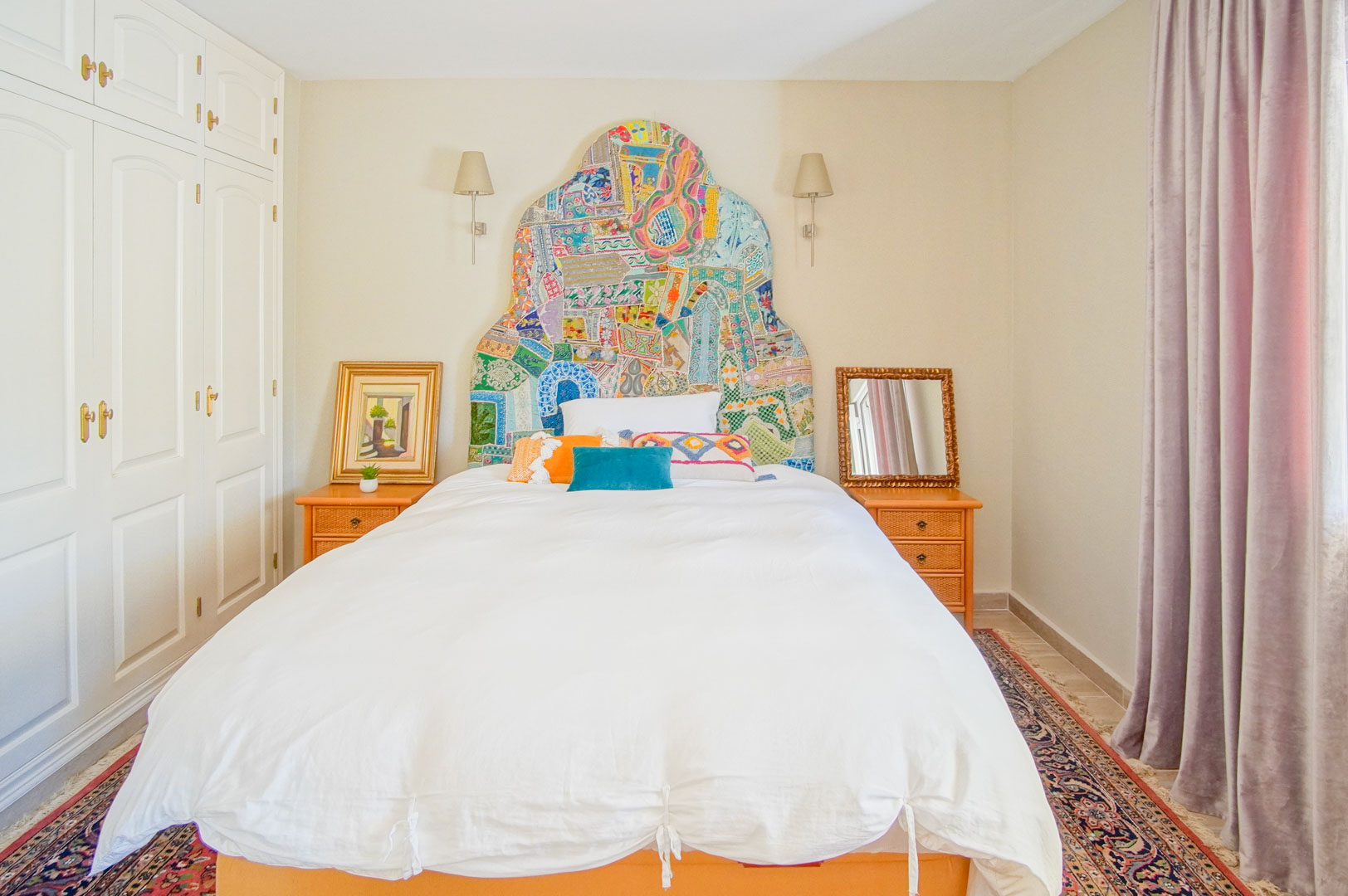Bedroom featuring a vibrant patchwork headboard, built-in white wardrobes, and wooden nightstands. Natural light illuminates the space, complemented by wall-mounted lamps.